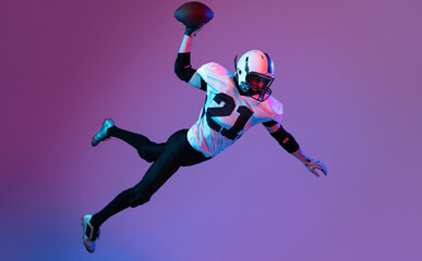Obraz na płótnie Canvas Portrait of american football player in motion, catching ball in a jump iisolated over purple background in neon light. Falling down