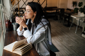 White woman in eyeglasses drinking coffee while reading book at cafe