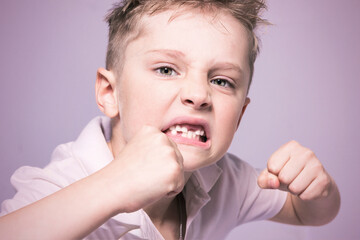 A preschooler with disheveled hair without one tooth opened his mouth and shows an aggressive pose with fists. Schoolboy shows strength. The blond boy grimaces at the camera