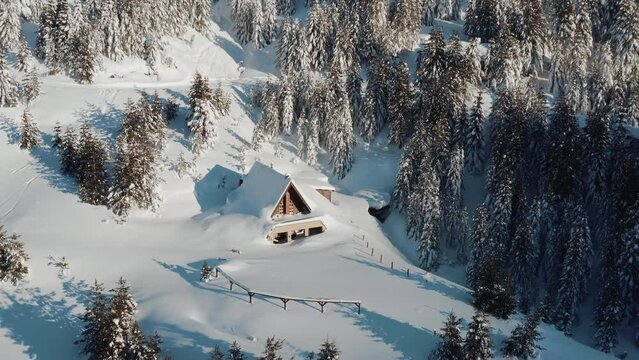 Drone shot of a small cottage house covered in snow on an icy winter morning on a sunny day. Winter fairytale in a remote forest.