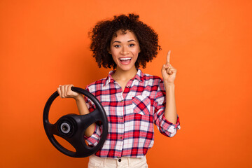 Portrait of attractive trendy cheerful girl holding steering wheel point up solution isolated over bright orange color background
