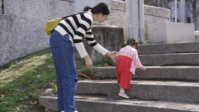 view from behind of Japanese mother supporting her young daughter climbing up stone stairs while taking a walk outside in an urban park on a sunny day.