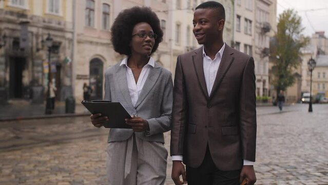 Beautiful woman with clipboard and handsome man with briefcase walking and talking on street. Two african colleagues in stylish formal wear posing outdoors.