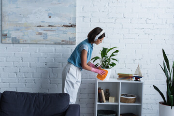 side view of woman in wireless headphones cleaning flowerpot on rack at home.