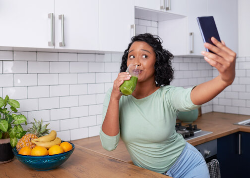 A young multiracial woman takes a selfie with her green smoothie in the kitchen