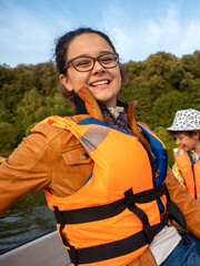 Beautiful brunette woman teen with glasses and in an orange life jacket rowing oars while sitting in a boat. Family walks at the boat station. Illuminated by sun glare