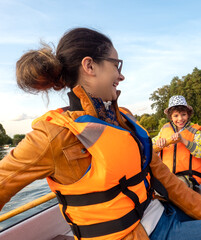 Beautiful brunette woman teen with glasses and in an orange life jacket rowing oars while sitting in a boat. Family walks at the boat station. Illuminated by sun glare
