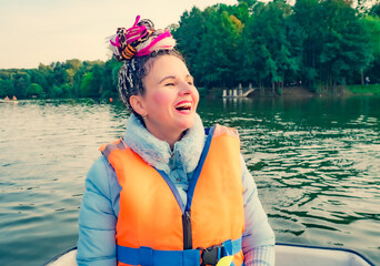 Middle age happy woman with fancy hairstyle wearing nautical lifejacket laughs and admires the views of beautiful autumn nature. Woman in a hat traveling on a boat