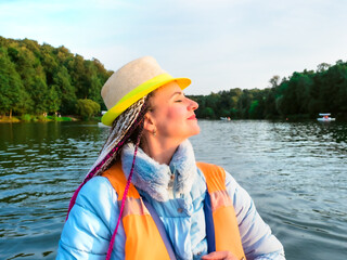 Middle age happy woman with fancy hairstyle wearing nautical lifejacket laughs and admires the views of beautiful autumn nature. Woman in a hat traveling on a boat