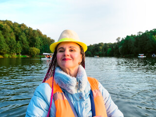 Middle age happy woman with fancy hairstyle wearing nautical lifejacket laughs and admires the views of beautiful autumn nature. Woman in a hat traveling on a boat