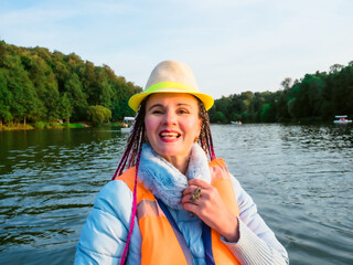 Middle age happy woman with fancy hairstyle wearing nautical lifejacket laughs and admires the views of beautiful autumn nature. Woman in a hat traveling on a boat