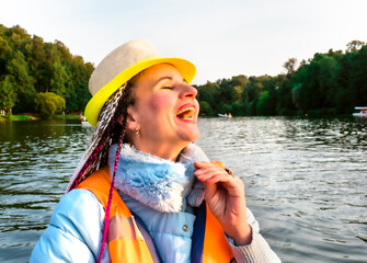 Middle age happy woman with fancy hairstyle wearing nautical lifejacket laughs and admires the views of beautiful autumn nature. Woman in a hat traveling on a boat