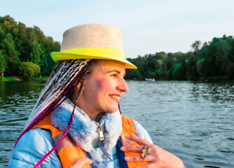 Middle age happy woman with fancy hairstyle wearing nautical lifejacket laughs and admires the views of beautiful autumn nature. Woman in a hat traveling on a boat