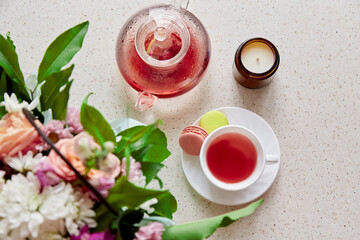 Obraz na płótnie Canvas Healthy raspberry natural tea glass teapot, macaroons, candle and bouquet of flowers. Herbal tea, top view. Coziness, self care, relaxing, wellness lifestyle.