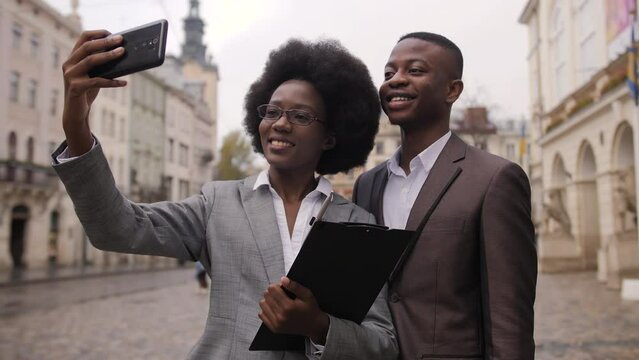 Successful afro american business people standing together on city street and taking selfie on smartphone. Young man and woman in formal clothes smiling and gesturing on camera.