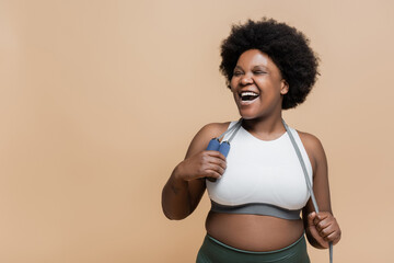 excited african american plus size woman in sportswear holding skipping rope isolated on beige.