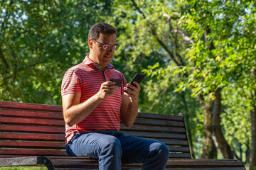 Obraz premium Happy millennial man using smartphone and credit card, sitting on wooden bench in sunny city park. Confident caucasian man purchasing online on cellphone