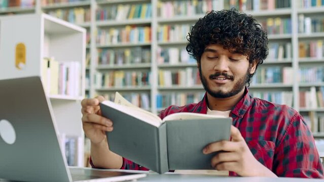 Positive smart arabian or indian college male student, sits at table with laptop in university library, preparing for exam, studying information, rereading his abstract, gain knowledge, smiles