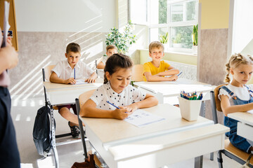 Five nice pupils are listening carefully to their tutor. Primary school kids sitting on desks and working in classroom. Education and school concept.