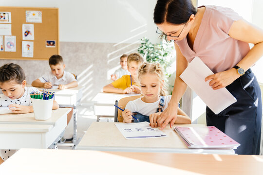 Nice pupils are listening carefully to their tutor. Primary school kids sitting on desks and working in classroom. Female teacher help cute little schoolgirl during lesson.