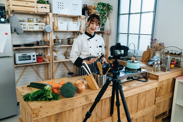 Asian woman chef is talking to the digital camera on tripod while filming and teaching cooking...