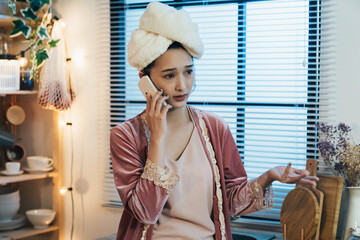 closeup confused asian woman wearing a nightgown with a towel on head is putting her palm up while having an unpleasant mobile phone talk at home.