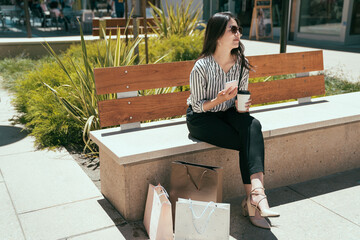 stylish Korean lady is using her mobile phone and enjoying sunlight while having a coffee break from shopping on the bench in an urban outdoor seating area.