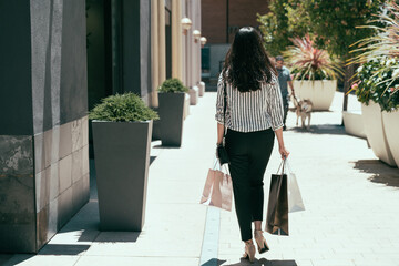 rear view and full length of an Asian woman walking alone on the sidewalk near a building with shopping bags on a sunny day in downtown area.