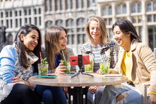 Four multi ethnic female friends, tourists or students sitting at a cafe terrace in the city center using mobile phone for a video call. High quality photo