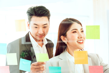 Young businesswoman office worker reading sticky notes on planning glass board, female corporate leader present project tasks on sticky notes organize work attaching stickers on scrum glass wall.