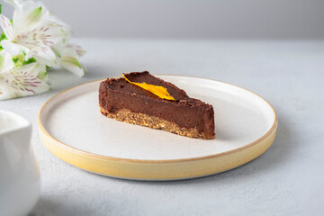 Slice of raw vegan chocolate cake on white plate with flowers on the background. Healthy vegan dessert, bake free, egg free, sugar free