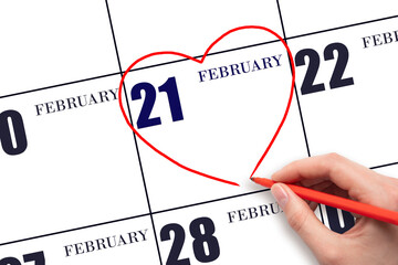 A woman's hand drawing a red heart shape on the calendar date of 21 February. Heart as a symbol of...