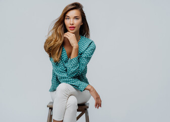 Indoor shot of lovely businesswoman sits on chair, holds chin, wears polka dot blouse and white...