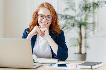 Pleased cheerful red haird female economist develops financial startup project, poses in office interior, works in business sphere, dressed in formal clothes, has happy expression, owns company