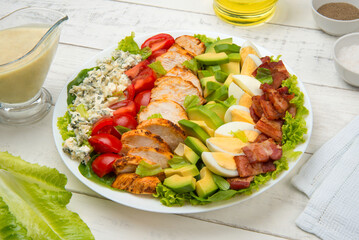 Cobb salad of romain lettuce, slices bacon, avocado, chicken, tomato, eggs, blue cheese in a white salad bowl on an old rustic white wooden table. - 527045374