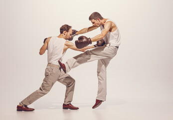 Portrait of two men playing, boxing in gloves isolated over grey studio background. Dynamic image...