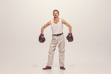 Portrait of muscular man in vintage clothes showing muscles in boxing gloves, posing isolated over grey studio background