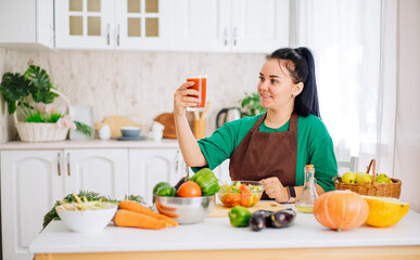 A young brunette woman in an apron is sitting at a table with fresh vegetables, looking at a glass of freshly squeezed homemade juice from fresh vegetables in a white kitchen.