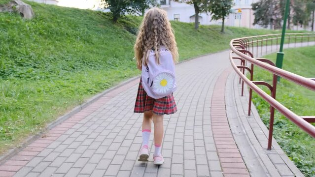 A happy girl with a backpack goes to school on the way. The child returns to school. A girl walks along a path in the park. Rear view