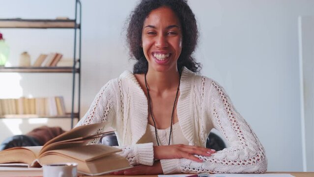 Young attractive African American woman teacher waving hand starting online lesson for distance school students with smile leads edu channel sits at table with open textbook. Education, training