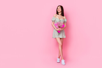Full length photo of cheerful dreamy lady wear green clothes looking empty space holding bouquet isolated pink color background