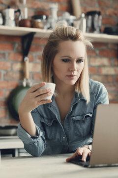 Image of young blond woman with short hair holding a cup of tea and working on a computer in the kitchen