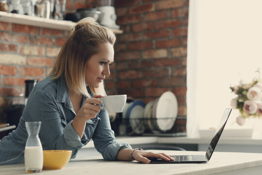 Picture of young woman with short hair holding a cup of coffee and working on computer in the kitchen