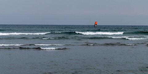 Grey sky over the Cold Sea. seascape. Windsurfing. A board with an orange sail. Surfing on the cold sea. Person surfing on sea waves