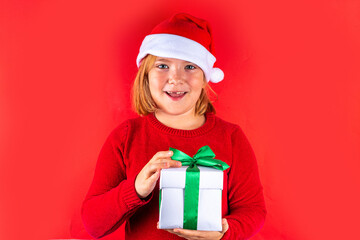 Christmas holiday background with funny blond girl. Little kid girl in red Christmas sweater and funny santa hat costume, with gift box on red background, Xmas greeting card, copy space