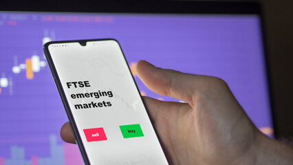 An investor's analyzing the FTSE emerging markets etf fund on screen. A phone shows the ETF's prices ftse London to invest