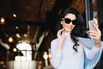 Smiling glamour woman in sunglasses, white blouse and jacket posing into camera of her smartphone while making selfie photographing herself. Happy elegant model posing in cafe using mobile phone