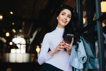 Dreamy stylish brunette lady with appealing appearance dressed elegantly holding mobile phone in hands watching photos remembering pleasant moments in her life. Woman with pensive expression