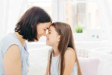 Close up shot of affectionate young mother and her little daughter touches noses and enjoy togetherness, pose against white background and bedroom interior, smile gladfully. Motherhood concept