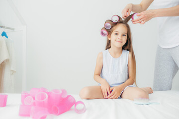Obraz na płótnie Canvas Horizontal shot of cute small daughter sits near her mother who winds curlers, pose against white background, prepares for festive event in kindergarten. Beautiful delighted kid has long hair